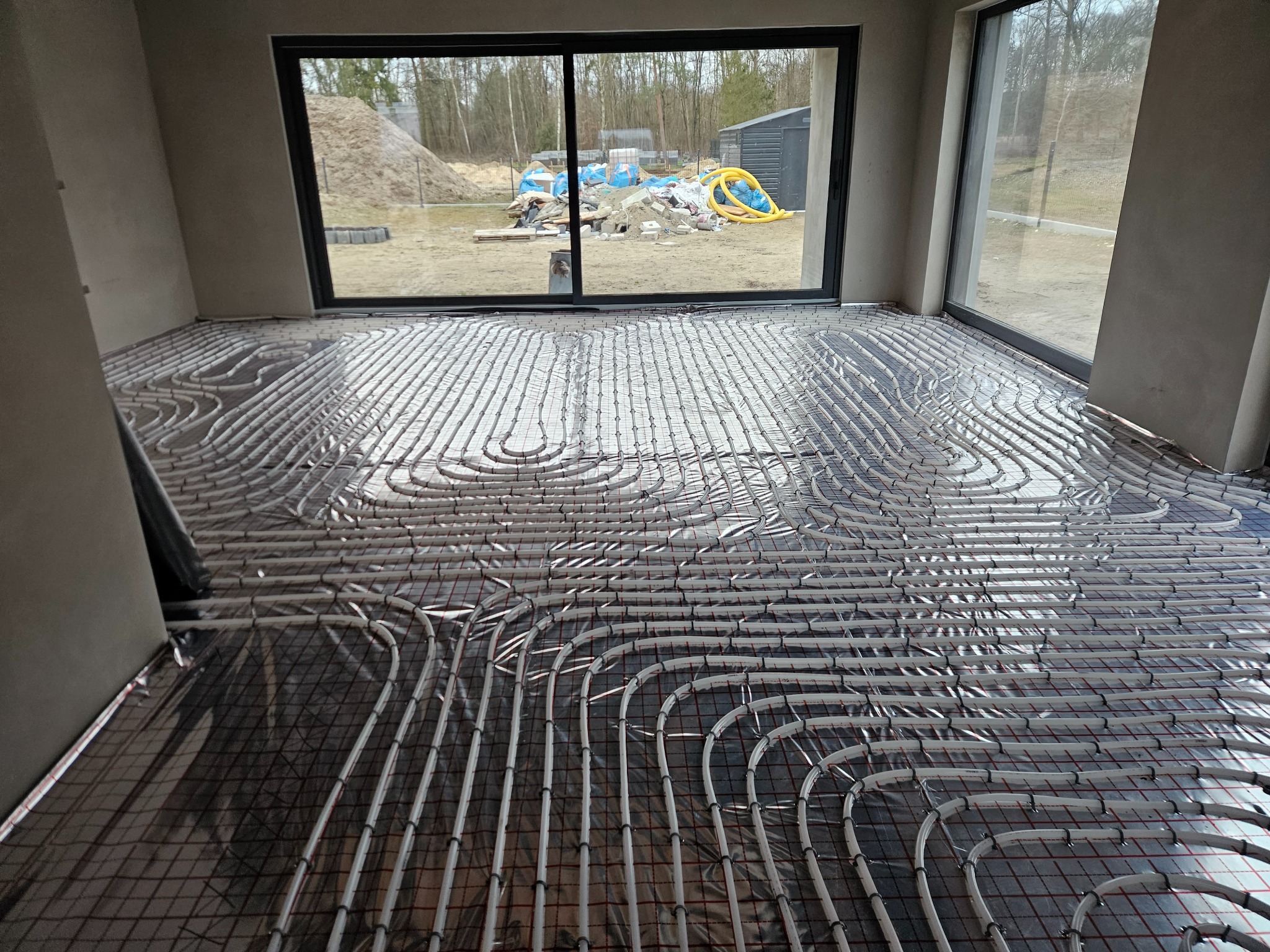 Rozłożona instalacja ogrzewania podłogowego na folii izolacyjnej w surowym pomieszczeniu z widokiem na plac budowy za oknem.