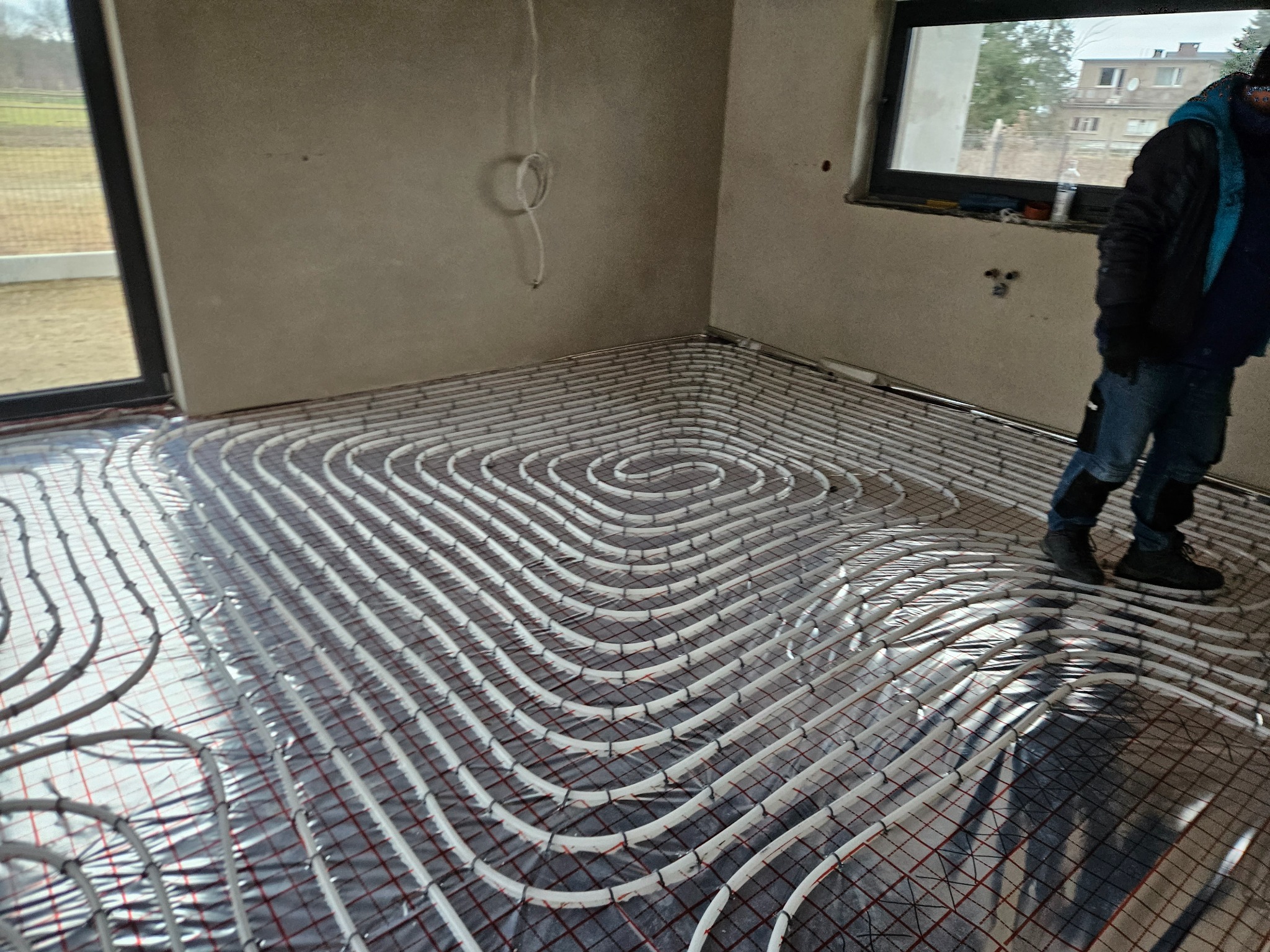 Układanie rur grzewczych na folii aluminiowej z siatką w surowym wnętrzu domu jednorodzinnego, widoczna osoba w roboczym ubraniu.