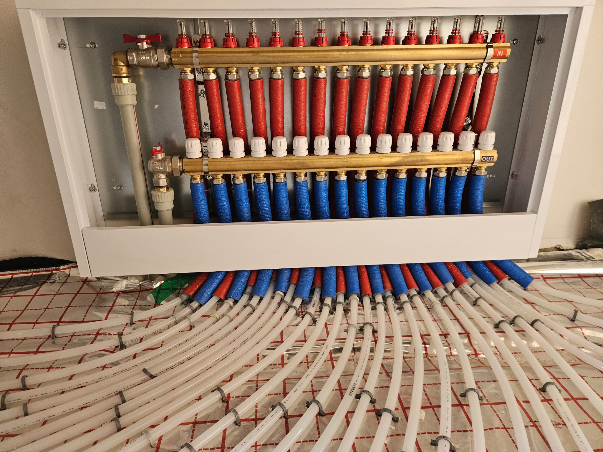 Rozdzielacz ogrzewania podłogowego z rurami rozprowadzonymi na folii izolacyjnej z nadrukowaną siatką, widoczne zawory i oznaczenia 'IN' i 'OUT'.
