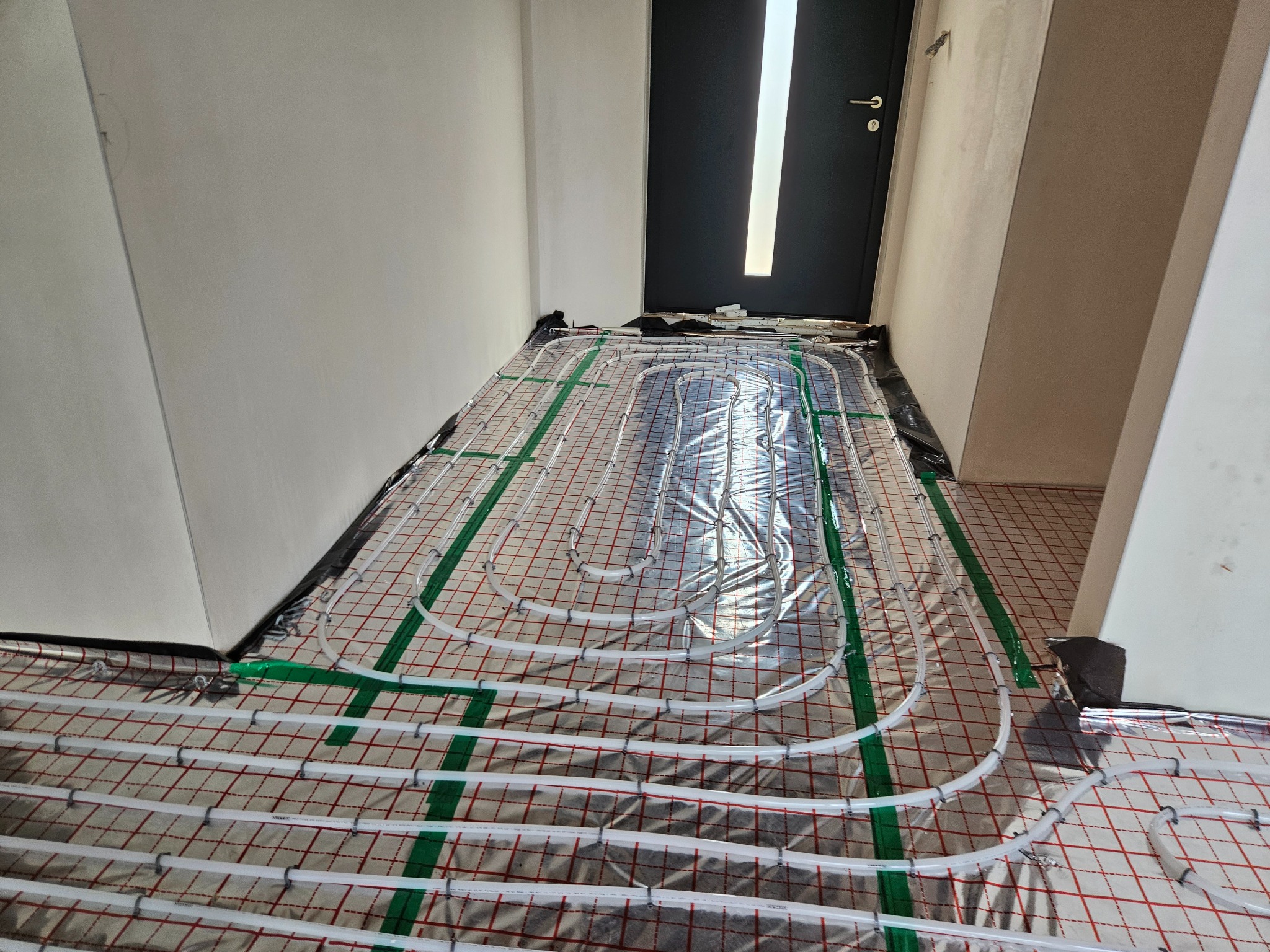 Rozłożona mata grzewcza podłogowa z rurkami na folii, widok na wejście z drzwiami w surowym wnętrzu budynku.