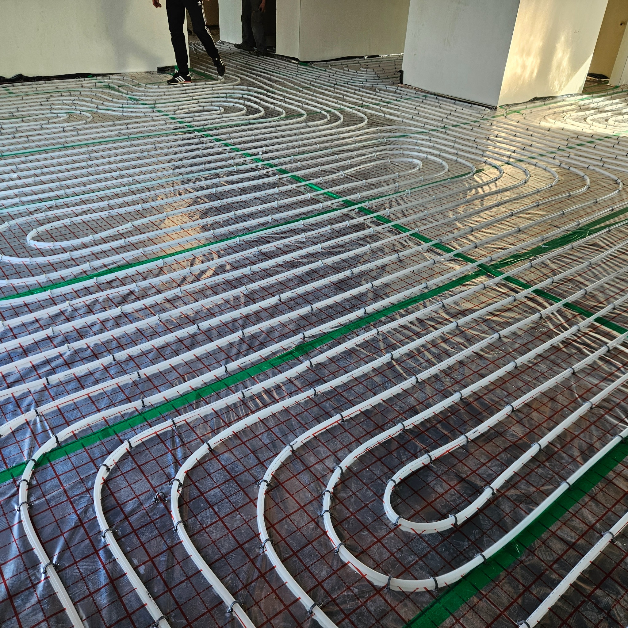 Rozłożona instalacja ogrzewania podłogowego na folii z siatką, widoczne białe rury grzewcze przymocowane czarnymi uchwytami i zielone paski mocujące, fragment osoby w tle.