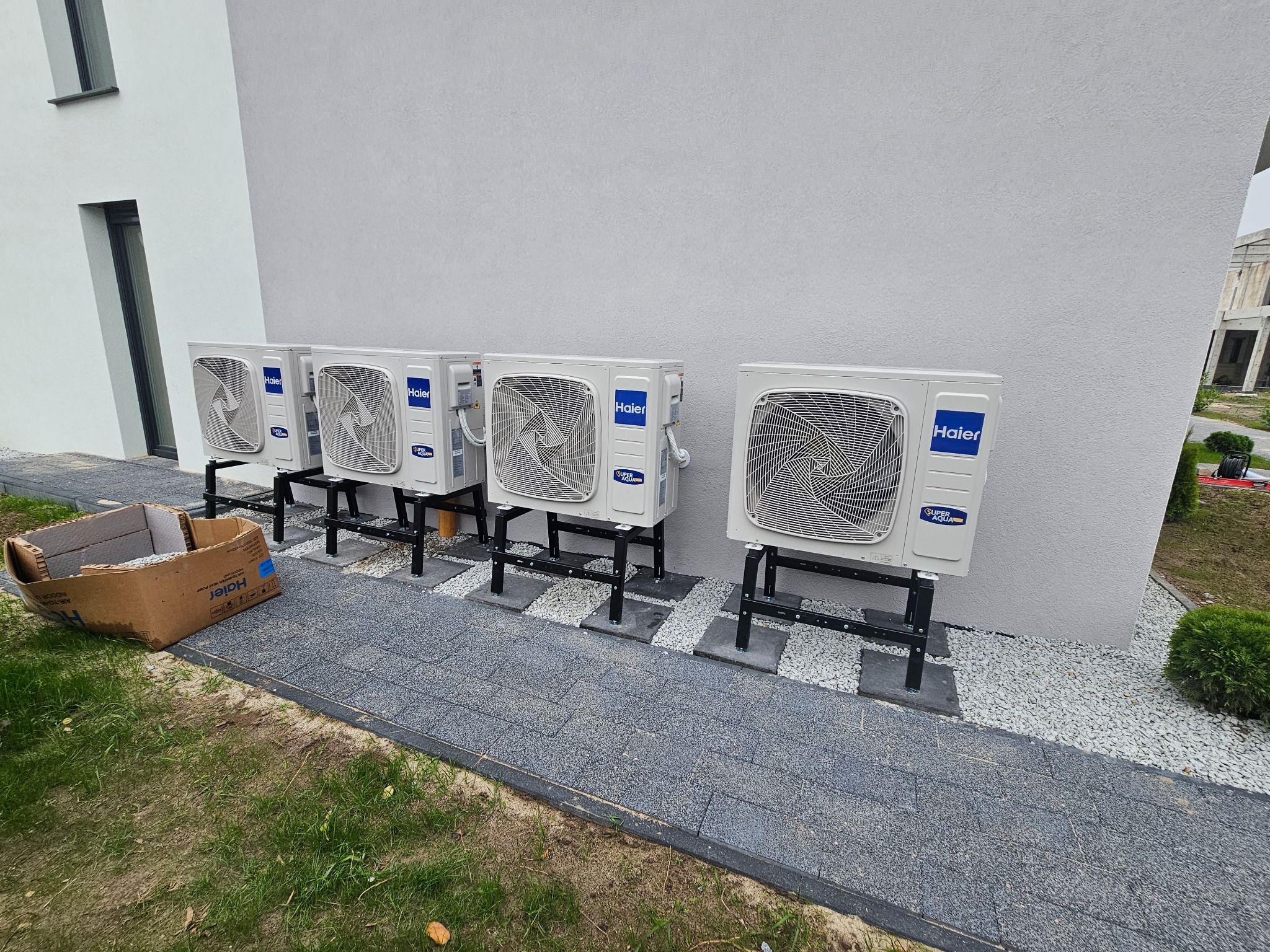 Rząd jednostek zewnętrznych klimatyzacji Haier zamontowanych na czarnych wspornikach wzdłuż ściany budynku, z kartonem po urządzeniu leżącym na trawie.