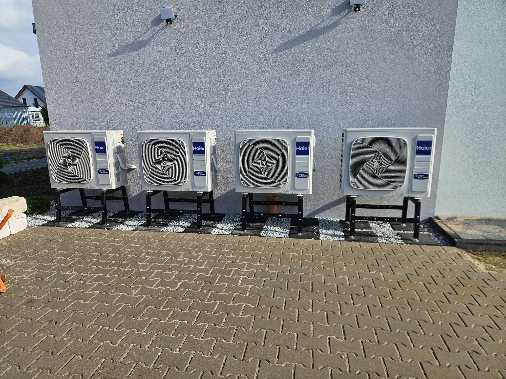 Cztery jednostki zewnętrzne klimatyzacji Haier zamontowane na czarnych wspornikach, ustawione rzędem na białym żwirze przy szarej ścianie budynku z widocznymi kamerami monitoringu.