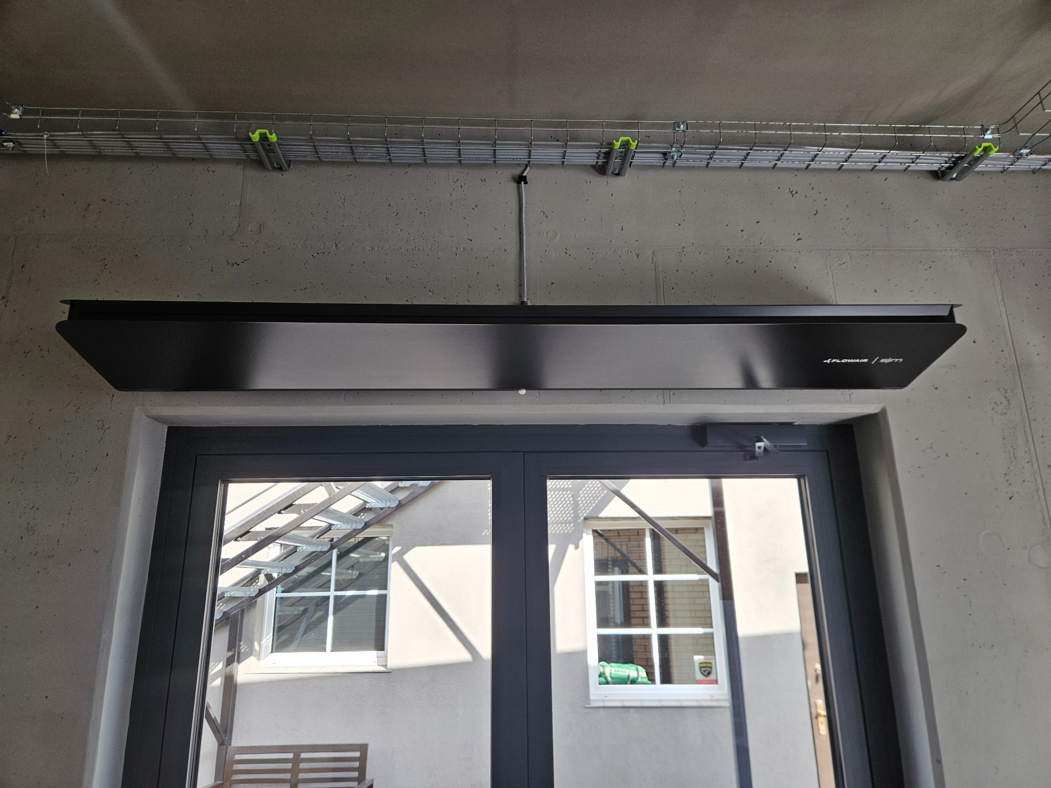Czarny, podwieszany system grzewczy Flowair Slim zamontowany nad drzwiami wejściowymi, widoczna instalacja kablowa na suficie z betonu architektonicznego.