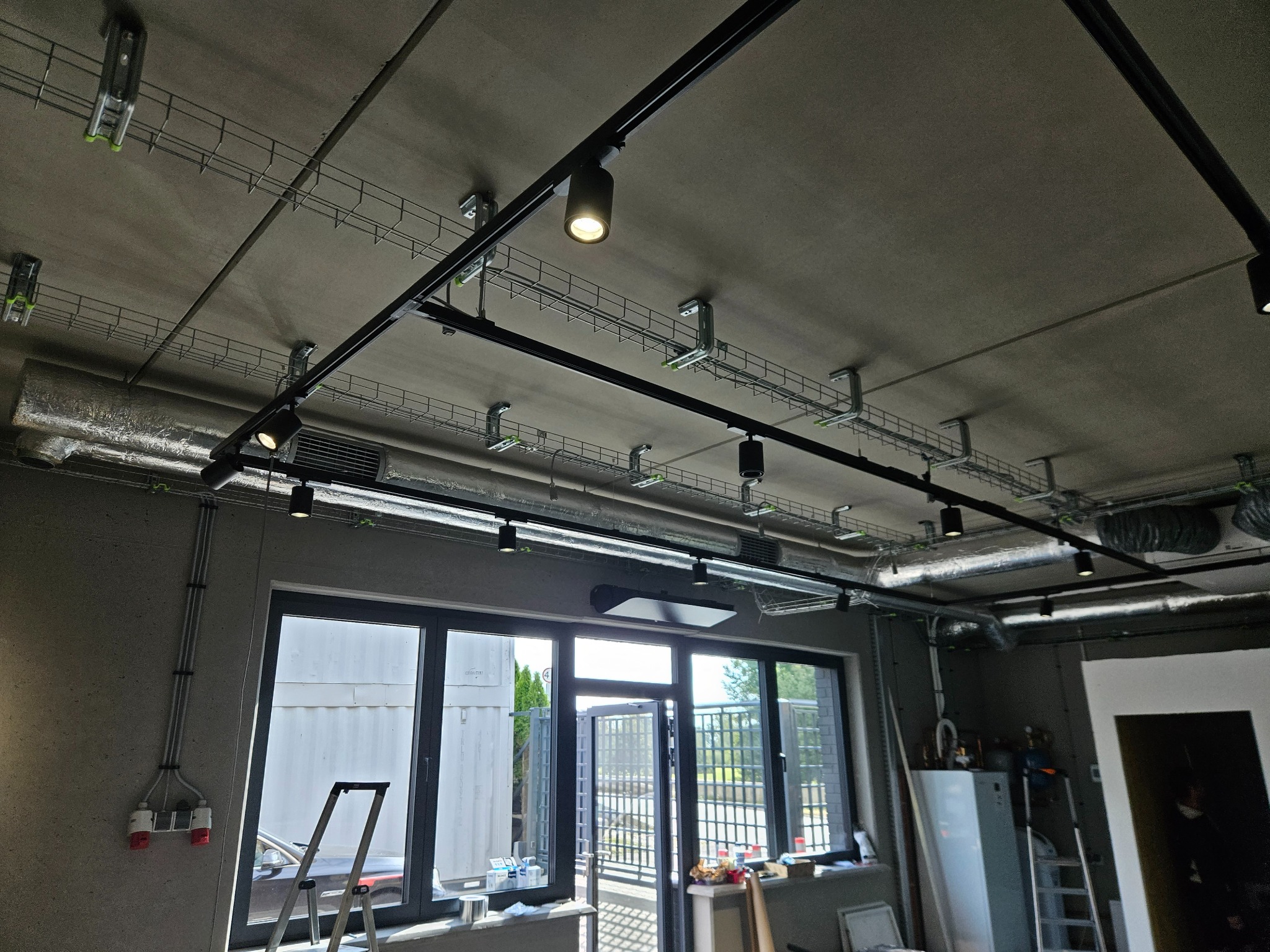 Widok sufitu w pomieszczeniu komercyjnym z widocznymi elementami instalacji wentylacyjnej, trasami kablowymi i oświetleniem punktowym. Na ścianie widoczne przewody elektryczne i okno z widokiem...