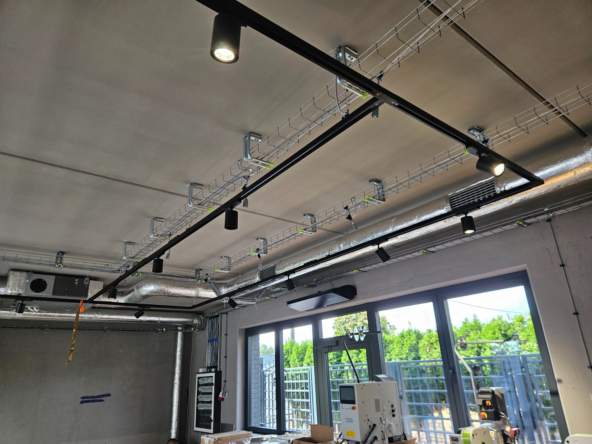 Widok na sufit z zamontowanymi korytami kablowymi, przewodami wentylacyjnymi i reflektorami punktowymi w nowoczesnym pomieszczeniu biurowym z dużymi oknami.