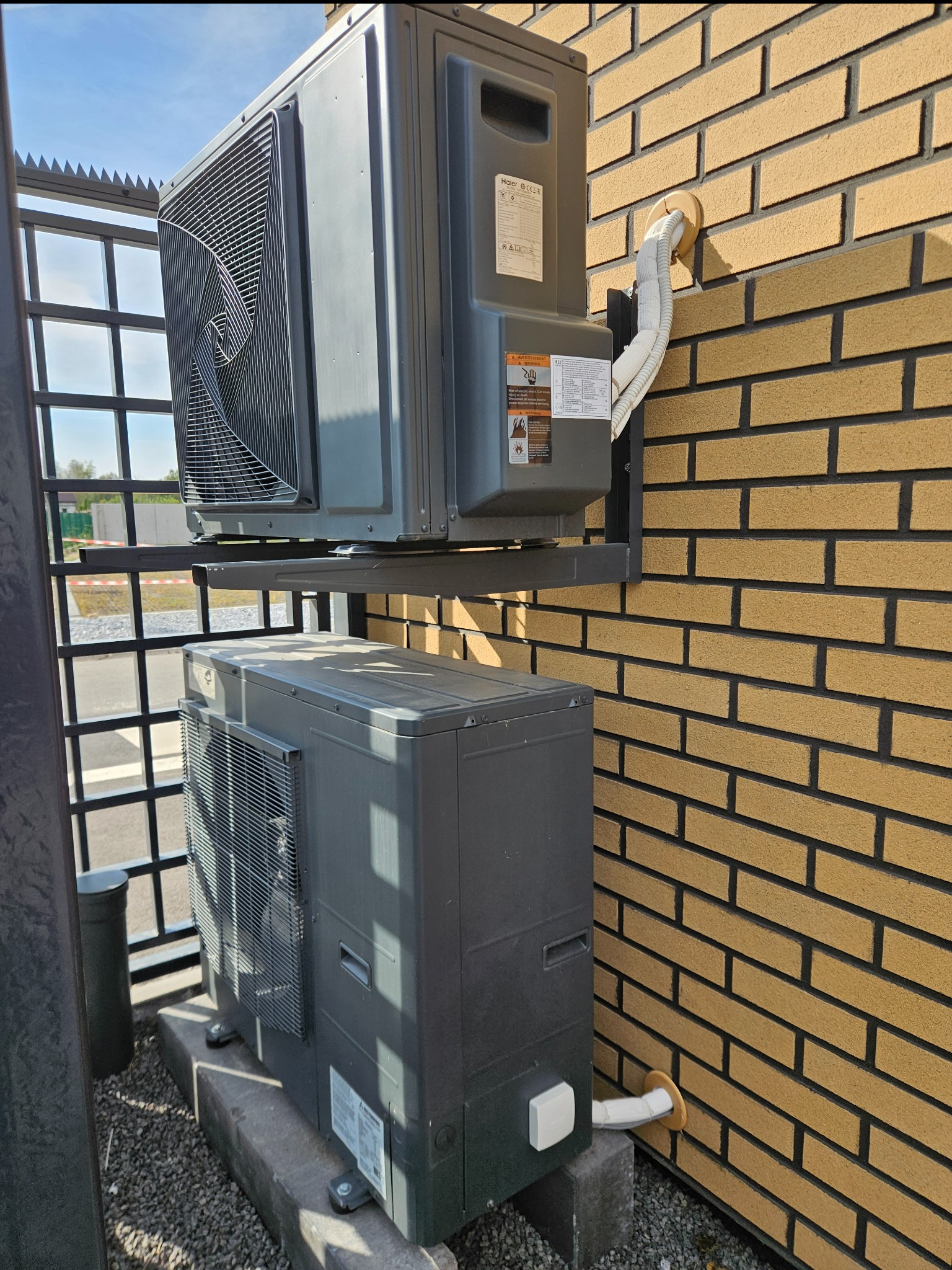 Dwa szare moduły urządzenia grzewczego zamontowane na zewnątrz budynku z cegły, jeden nad drugim, połączone rurami, widoczne elementy montażowe i otoczenie budynku.