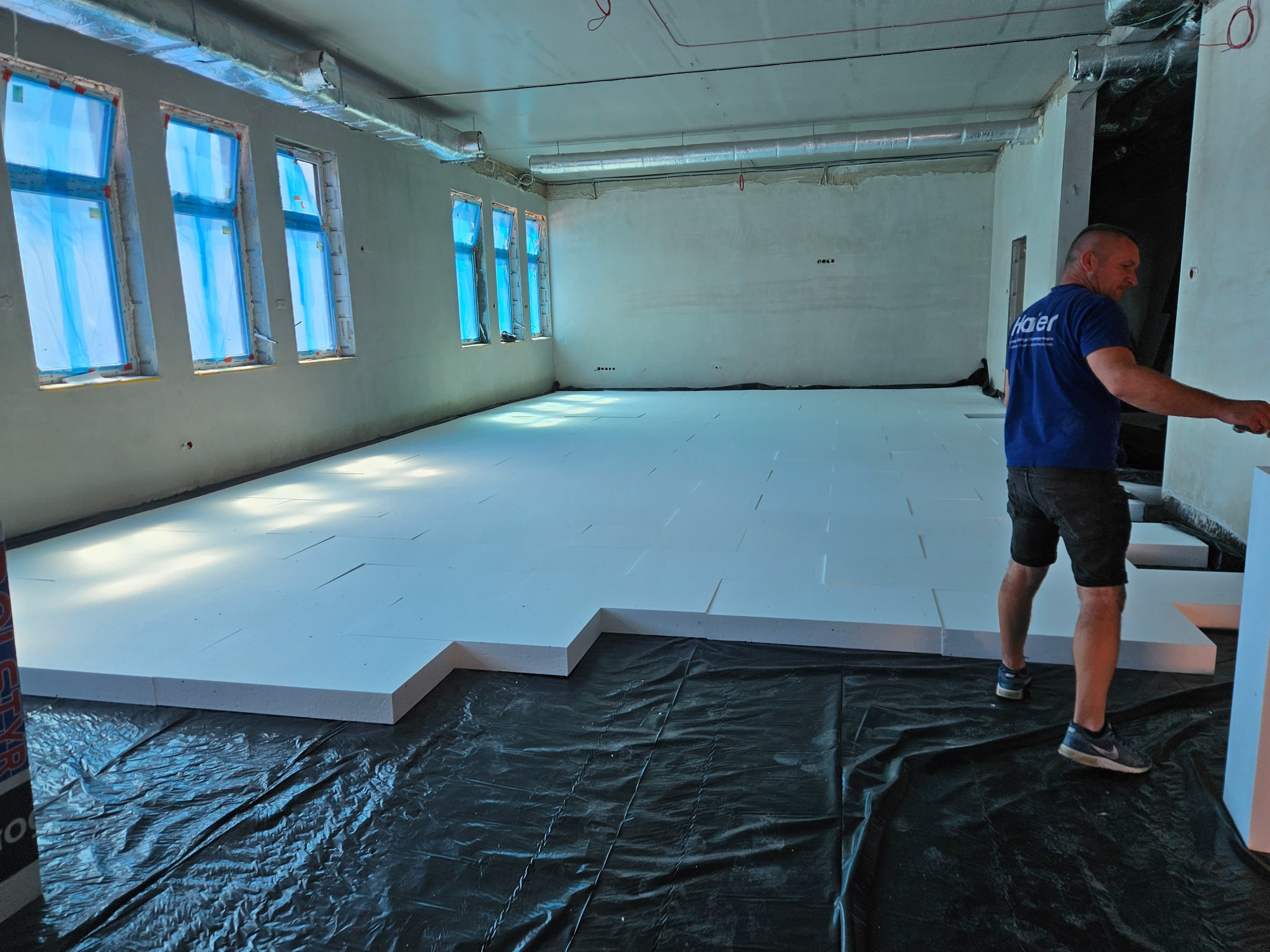 Układanie białych płyt izolacyjnych na czarnej folii w dużym pomieszczeniu z oknami zabezpieczonymi folią, widoczna instalacja wentylacyjna pod sufitem oraz pracownik w niebieskiej koszulce z logo...