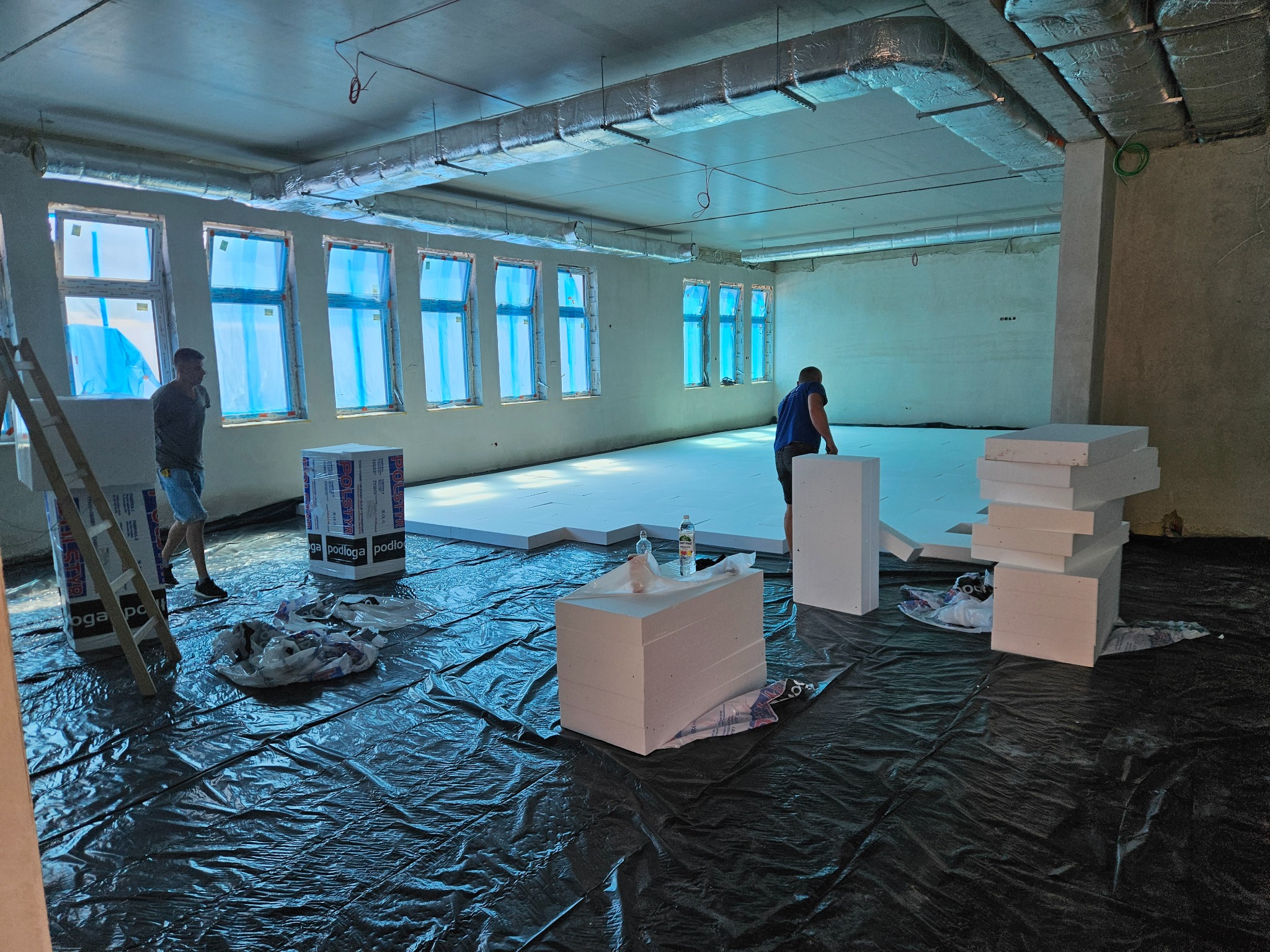 Montaż izolacji podłogowej z białych płyt styropianowych w surowym pomieszczeniu z oknami zabezpieczonymi folią, widoczne elementy wentylacji pod sufitem i czarna folia ochronna na podłodze.