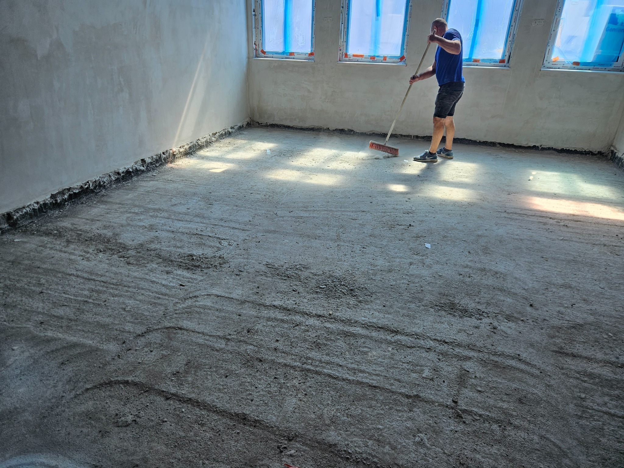 Mężczyzna zamiata podłogę w surowym stanie w pomieszczeniu z trzema oknami zabezpieczonymi folią malarską, przygotowanie do dalszych prac wykończeniowych.