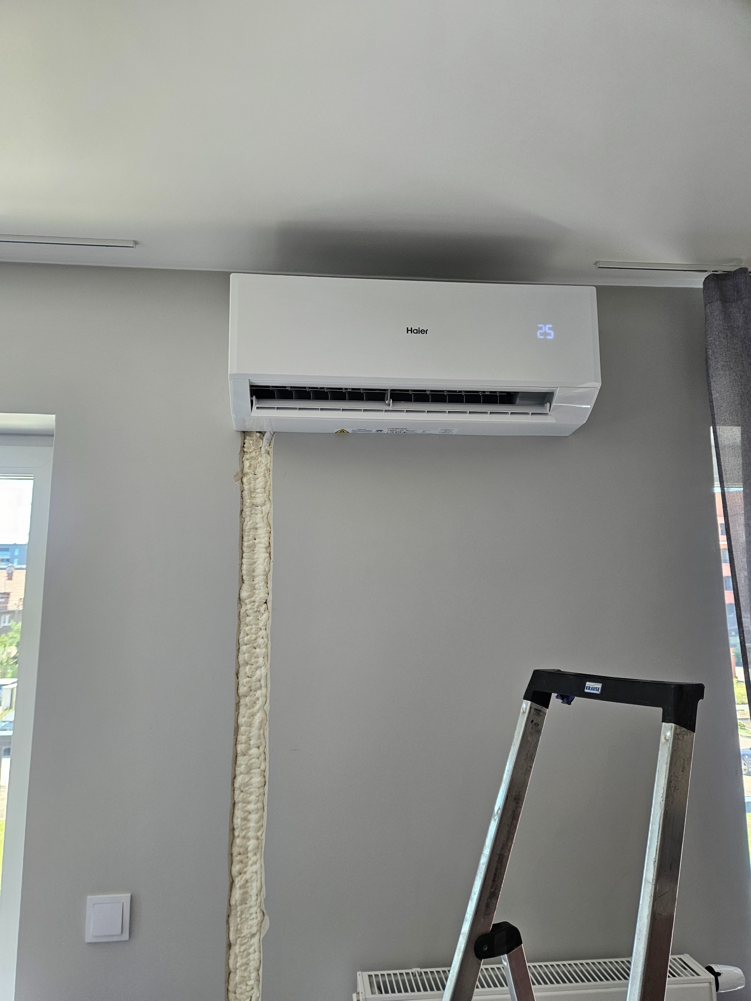 Biały klimatyzator Haier zamontowany na szarej ścianie, widoczna piana montażowa uszczelniająca otwór, drabina Krause w pomieszczeniu.
