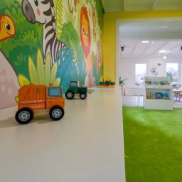 Waldemar Siębor Centrum opieki do lat 3 - Wnętrze pokoju zabaw z kolorową tapetą ze zwierzętami, na białym blacie stoją drewniane zabawki: pomarańczowa śmieciarka i zielony traktor, w tle widoczny zielony dywan i białe regały z zabawkami.