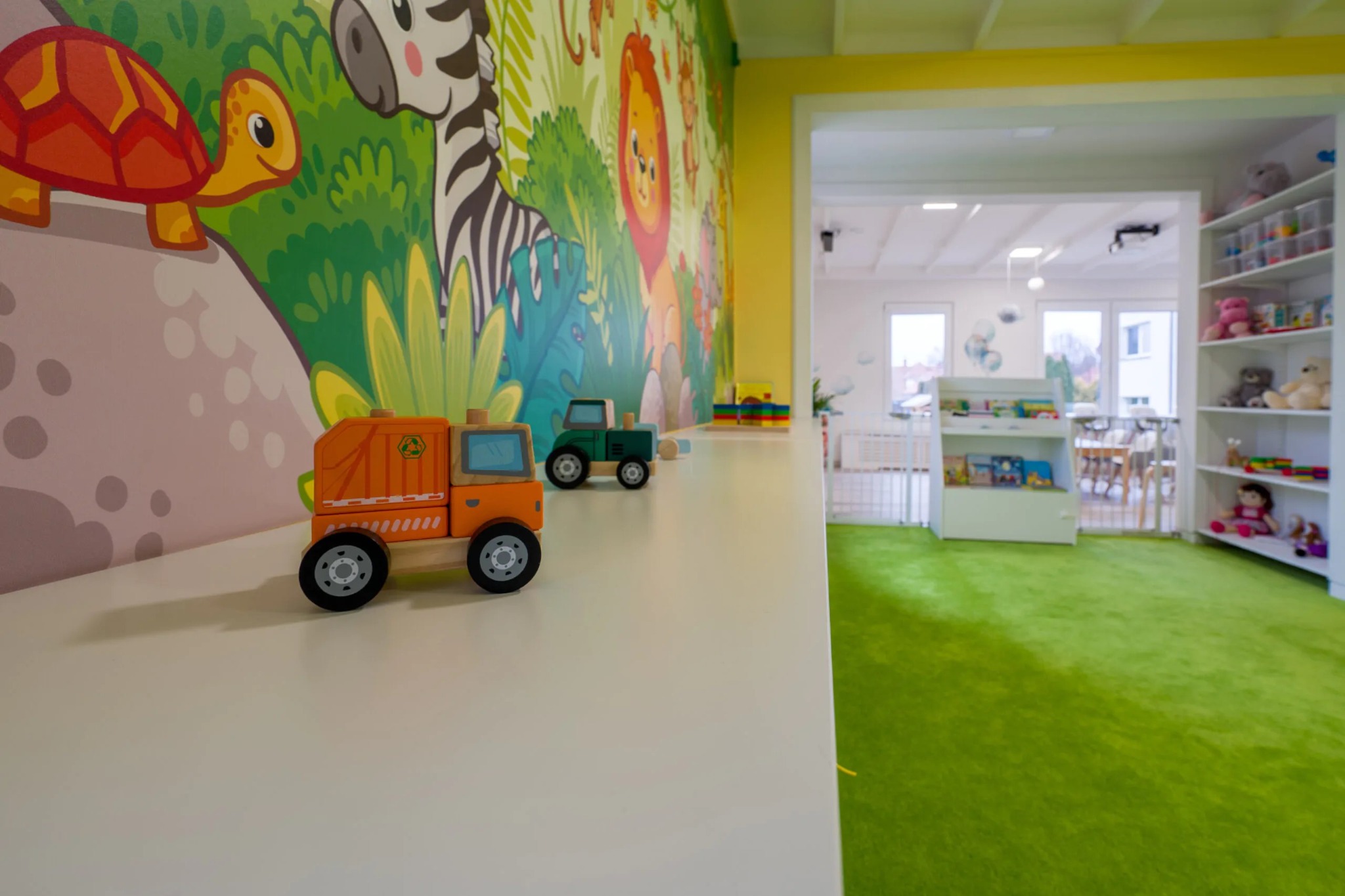 Wnętrze pokoju zabaw z kolorową tapetą ze zwierzętami, na białym blacie stoją drewniane zabawki: pomarańczowa śmieciarka i zielony traktor, w tle widoczny zielony dywan i białe regały z zabawkami.