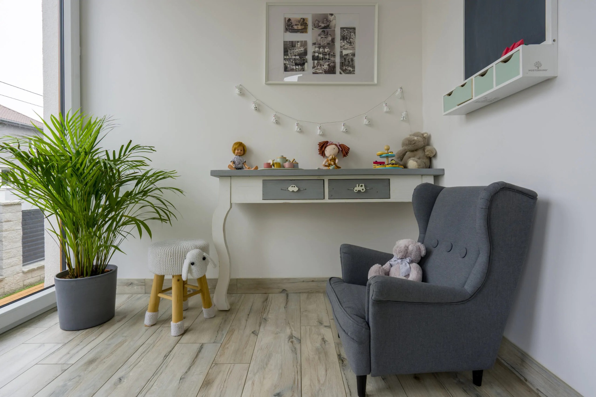 Jasny pokój dziecięcy z szarym fotelem uszakiem, biurkiem z szufladami z motywem aut, tablicą kredową i rośliną doniczkową w szarej osłonce.