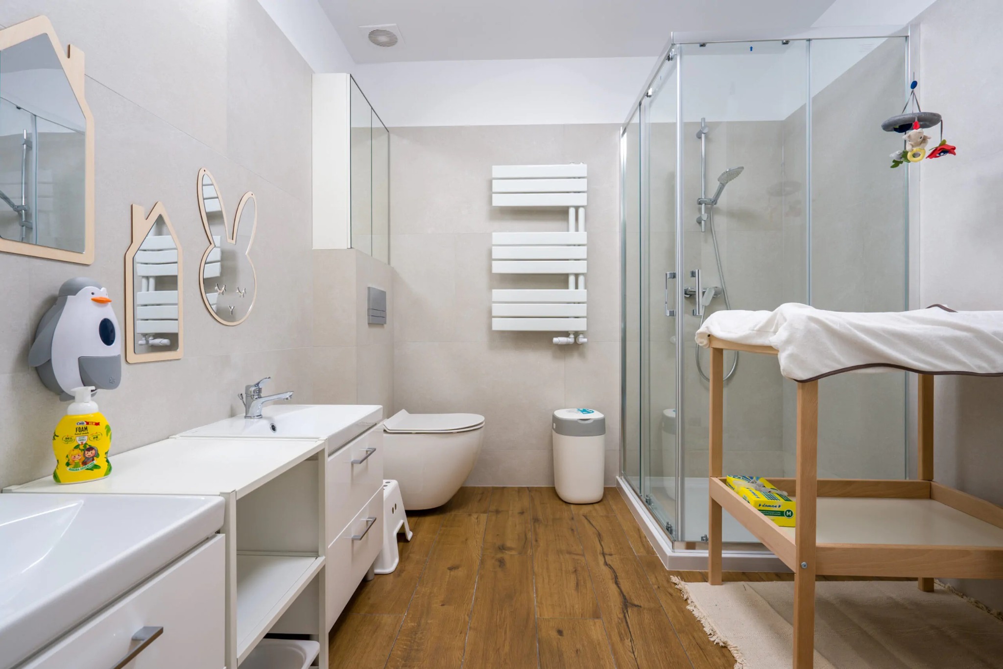 Jasna łazienka z drewnianą podłogą, wyposażona w przewijak z białym ręcznikiem, kabinę prysznicową, umywalki z dozownikiem mydła w kształcie pingwina i dekoracyjne lustra w kształcie domku i królika.
