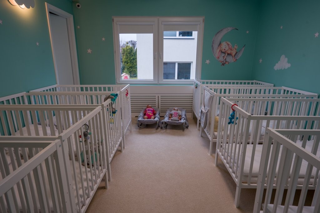 Pokój z wieloma białymi łóżeczkami dla niemowląt, dekoracjami ściennymi z motywem księżyca i gwiazd oraz dwoma leżaczkami z lalkami pośrodku.