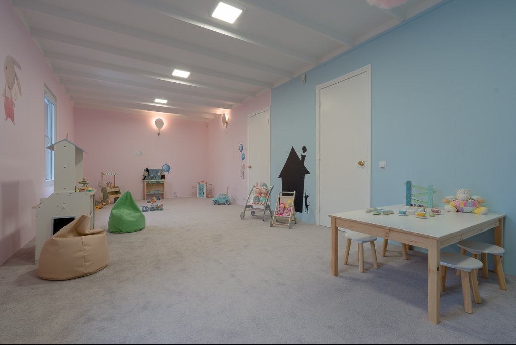Jasny pokój zabaw z pastelowymi ścianami, wyposażony w stolik z krzesełkami, zabawki, domki dla dzieci i pufy.