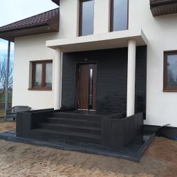Błażej Kaźmierczyk - Nowoczesne wejście do domu z czarnymi, granitowymi schodami i podestem, kontrastujące z białą elewacją i brązowymi oknami, teren wokół w trakcie prac wykończeniowych, widoczny piasek.