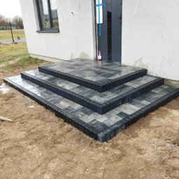 Błażej Kaźmierczyk - Schody wejściowe do domu wyłożone kostką brukową w odcieniach szarości i czerni, widoczne ślady wilgoci na kostce, surowy teren wokół schodów, drzwi wejściowe zabezpieczone folią.