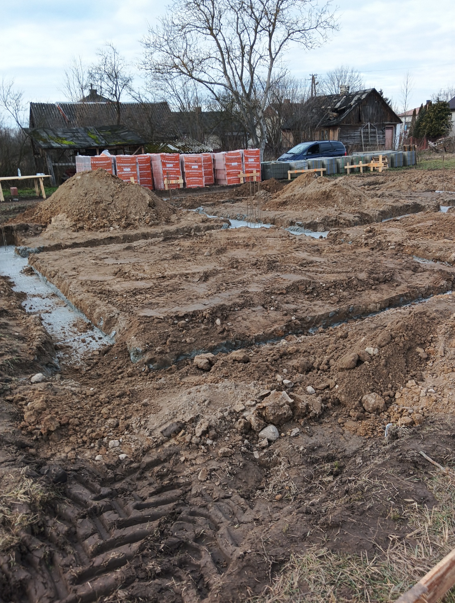Przygotowany wykop pod fundamenty z częściowo wylaną ławą fundamentową w Opocznie, widoczne ślady opon ciężkiego sprzętu i sterta cegieł w tle.
