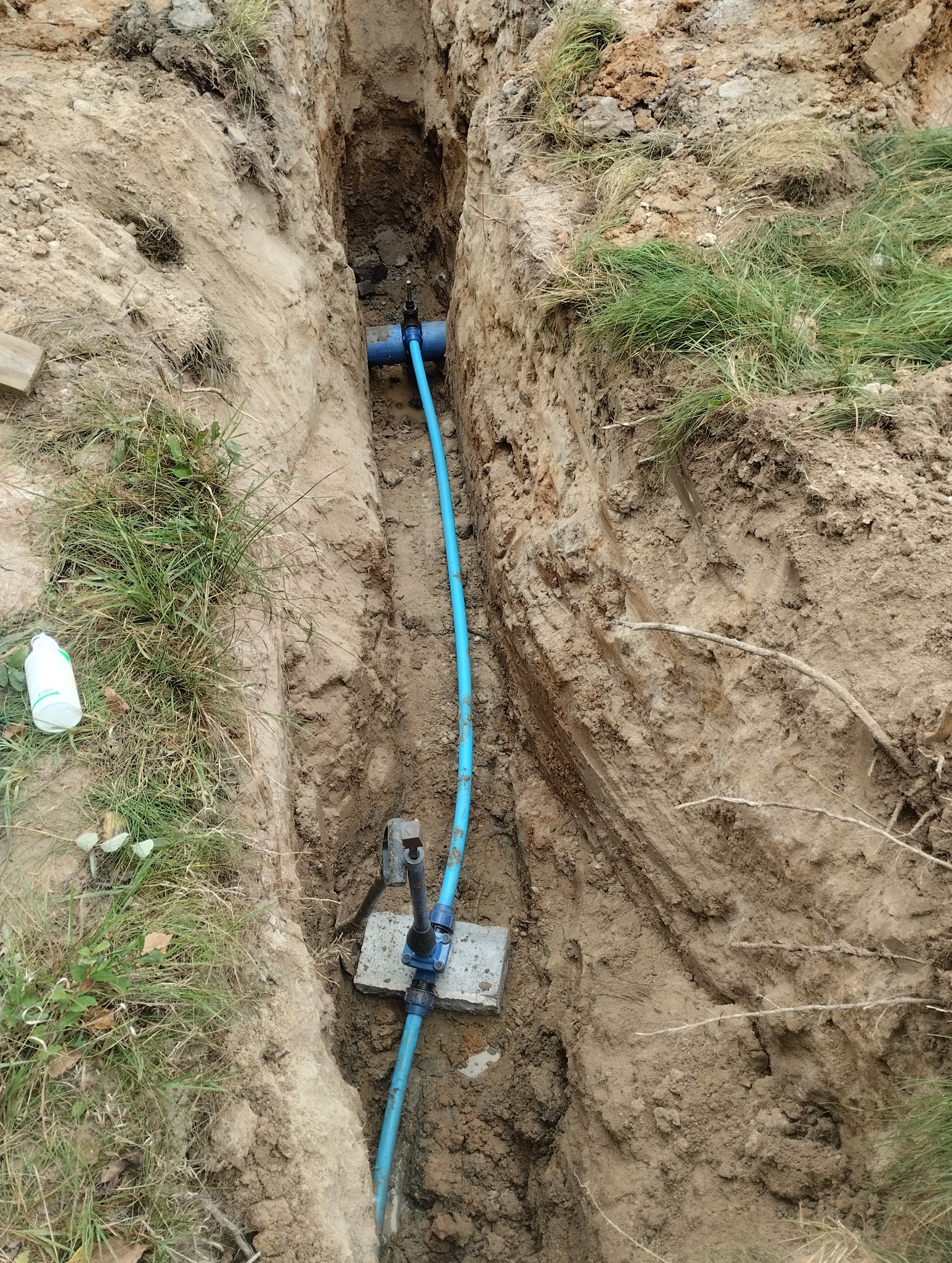 Wykop w ziemi z widoczną, niebieską rurą wodociągową z zamontowaną armaturą na betonowej podstawie, trawa na krawędzi wykopu, butelka w tle.
