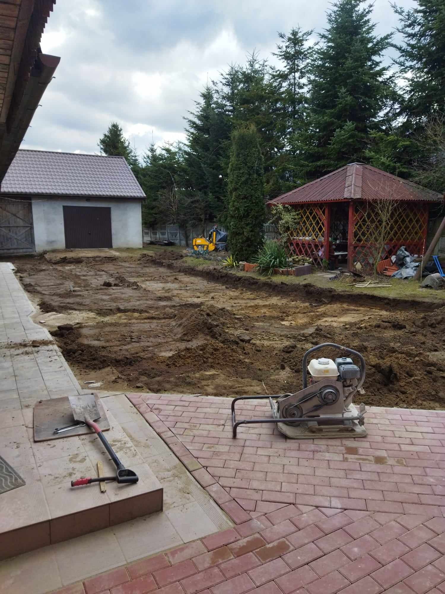 Przygotowany teren pod budowę z widoczną ubijarką wibracyjną, szpadlem i łopatą na kostce brukowej, w tle budynek gospodarczy, altana i drzewa.