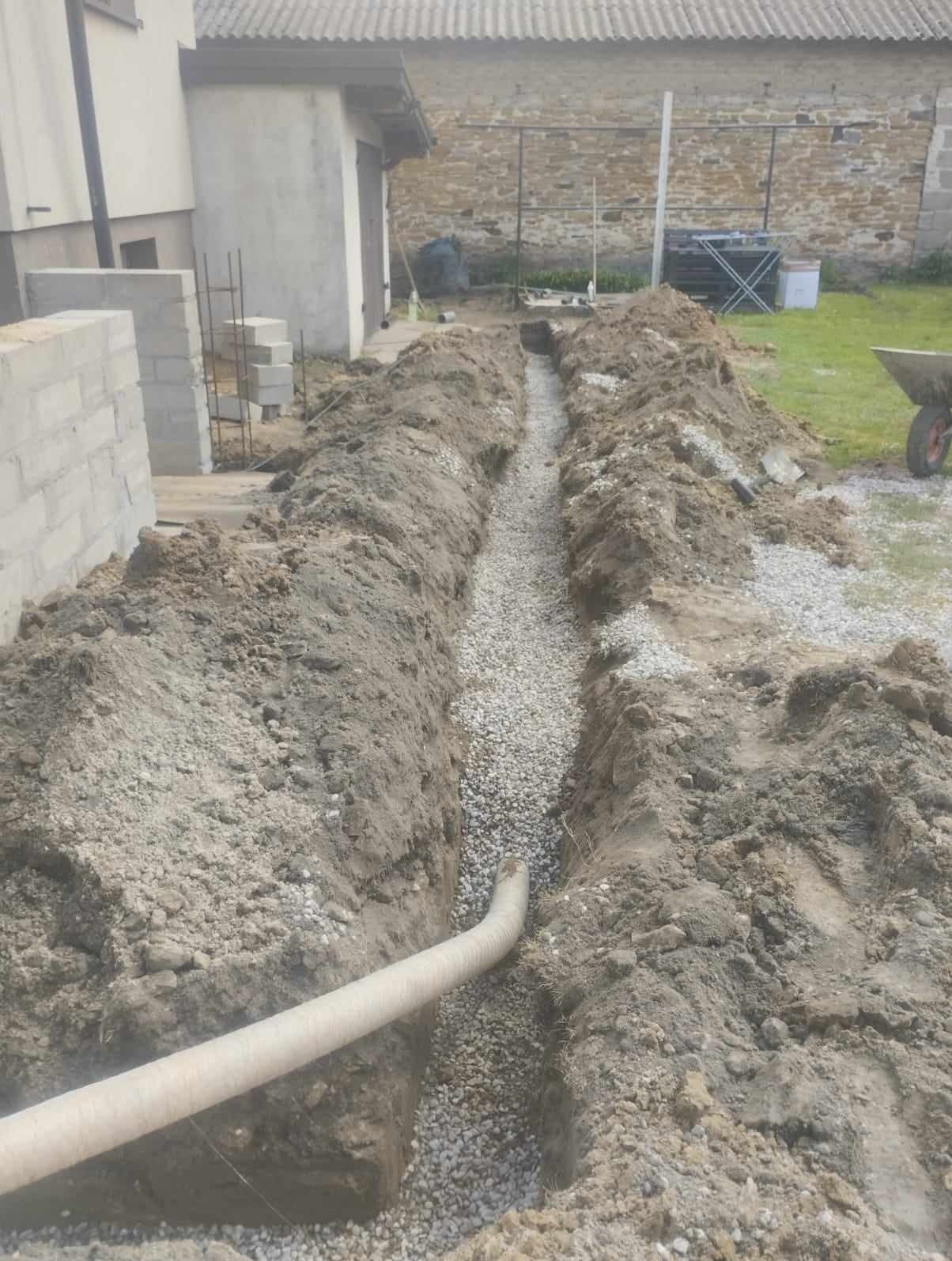 Wykopany wąski rów wypełniony drobnym żwirem, z widocznym fragmentem rury w dolnej części, na tle budynku gospodarczego i trawnika.