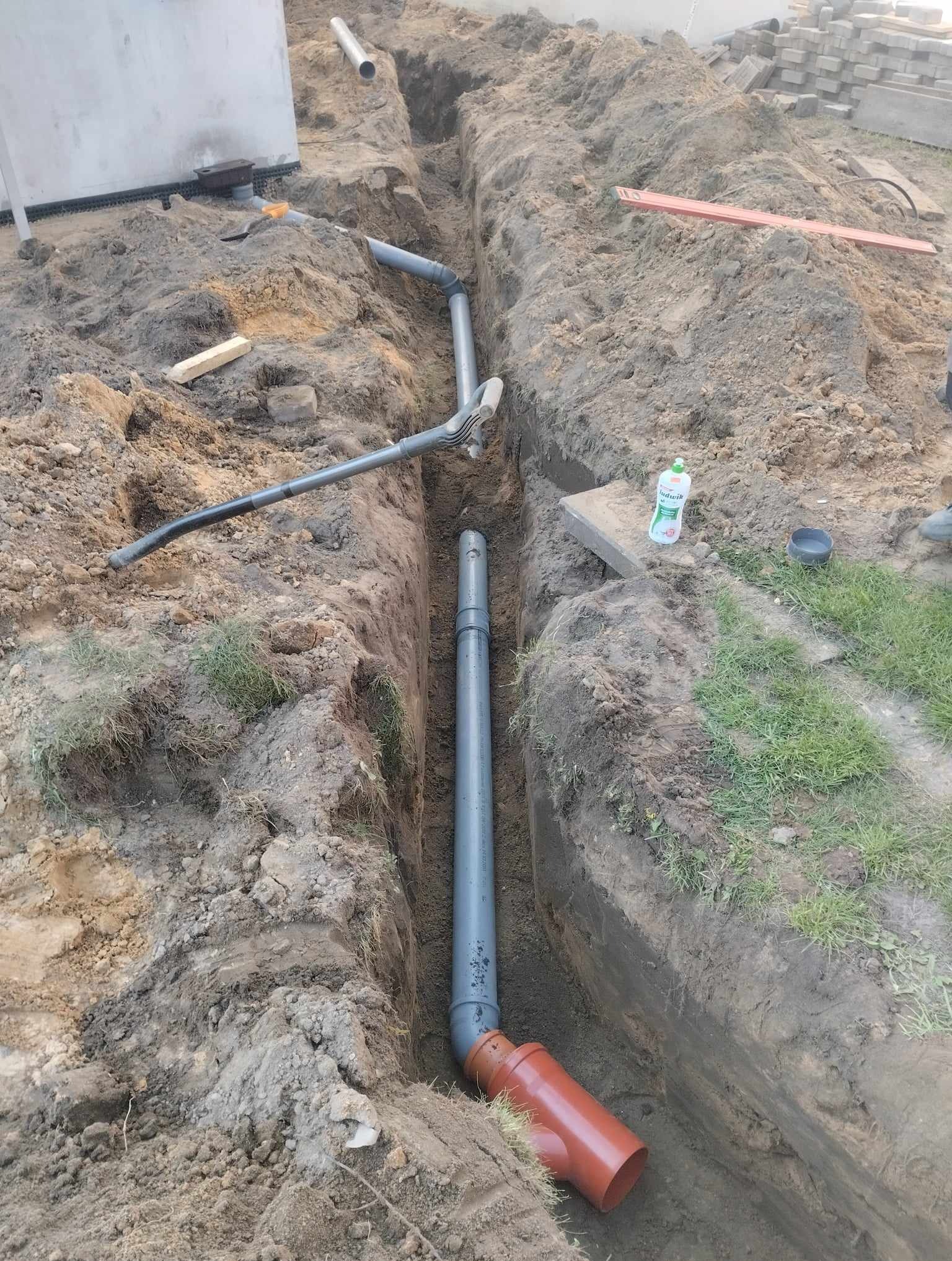 Głęboki wykop w ziemi z ułożonymi rurami kanalizacyjnymi z kształtkami, widoczny klucz do rur, poziomica i butelka płynu do naczyń.