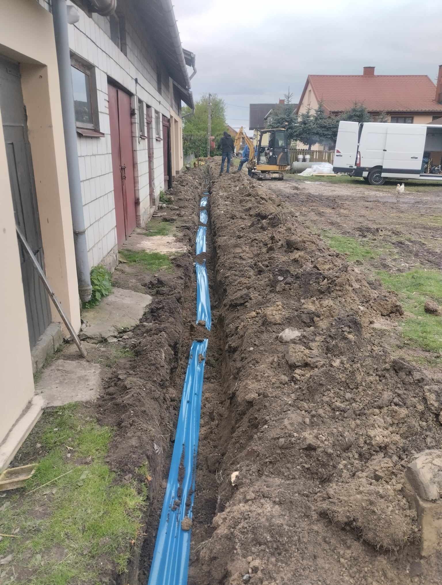Długi wykop w ziemi wzdłuż budynku gospodarczego, z widocznymi niebieskimi rurami ułożonymi na dnie, pracownicy i mała koparka w tle, teren nierówny, w tle budynki mieszkalne i biały samochód...