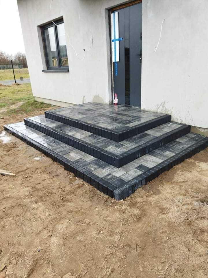 Schody wejściowe do domu wyłożone kostką brukową w odcieniach szarości i czerni, widoczne ślady wilgoci na kostce, surowy teren wokół schodów, drzwi wejściowe zabezpieczone folią.