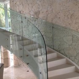 PROGRES SPÓŁKA Z OGRANICZONĄ ODPOWIEDZIALNOŚCIĄ - Eleganckie schody z białymi stopniami i zakrzywioną szklaną balustradą z metalowymi mocowaniami na tle ściany z naturalnego kamienia.