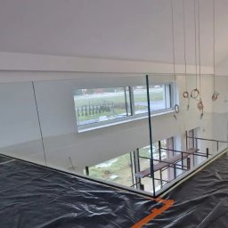 PROGRES SPÓŁKA Z OGRANICZONĄ ODPOWIEDZIALNOŚCIĄ - Szklana barierka zamontowana na antresoli, widoczne zabezpieczenia podłoża folią malarską, w tle okno z widokiem na zewnątrz i rusztowanie na dole.