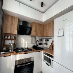 PROGRES SPÓŁKA Z OGRANICZONĄ ODPOWIEDZIALNOŚCIĄ - Kompaktowa kuchnia w nowoczesnym stylu z białymi i drewnopodobnymi szafkami, czarnym zlewem i armaturą, oraz białymi płytkami na ścianie, oświetlona trzema punktowymi lampami sufitowymi.