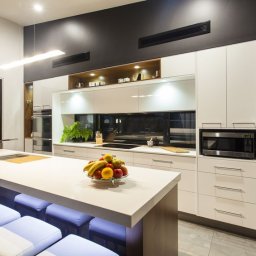 PROGRES SPÓŁKA Z OGRANICZONĄ ODPOWIEDZIALNOŚCIĄ - Nowoczesna kuchnia z białymi szafkami, drewnianymi akcentami i wyspą z podświetlanymi stołkami barowymi. Na blacie wyspy misa z owocami. Ciemnoszara ściana z wbudowanymi kratkami wentylacyjnymi.