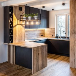 PROGRES SPÓŁKA Z OGRANICZONĄ ODPOWIEDZIALNOŚCIĄ - Nowoczesna kuchnia z czarnymi szafkami i blatem z drewna o naturalnym odcieniu, oświetlona wiszącą lampą z roślinami w doniczkach, z widocznym barkiem z pionowymi listwami.
