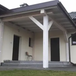 Zadaszone wejście do domu z drewnianą konstrukcją wspartą na białych słupach, schody wyłożone szarymi płytami, widok na dwie pary drzwi wejściowych i krzesło stojące obok.