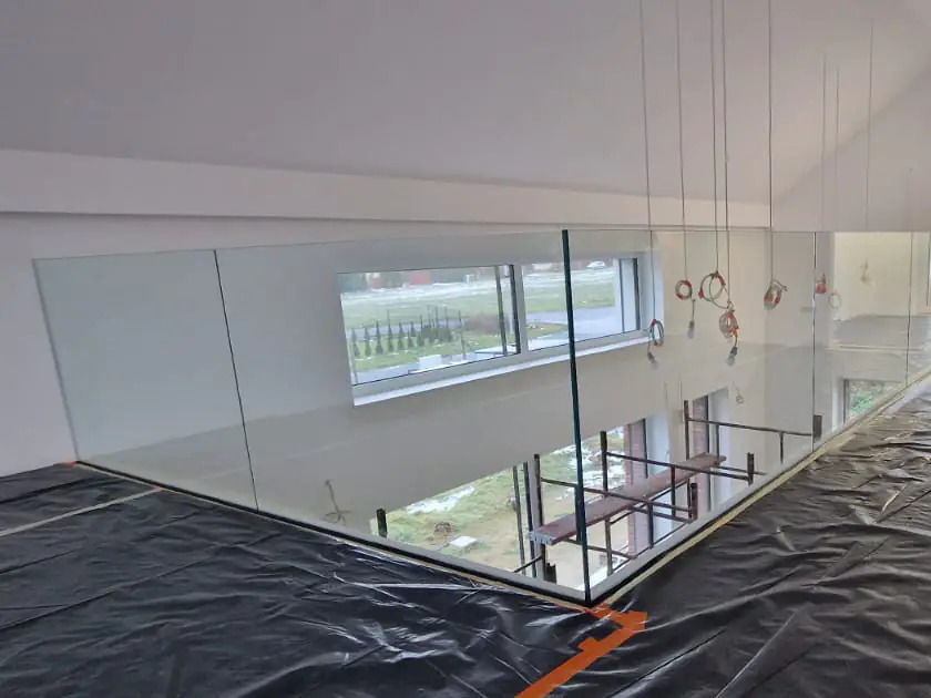 Szklana barierka zamontowana na antresoli, widoczne zabezpieczenia podłoża folią malarską, w tle okno z widokiem na zewnątrz i rusztowanie na dole.