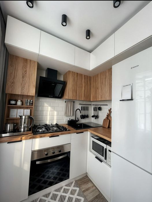 Kompaktowa kuchnia w nowoczesnym stylu z białymi i drewnopodobnymi szafkami, czarnym zlewem i armaturą, oraz białymi płytkami na ścianie, oświetlona trzema punktowymi lampami sufitowymi.