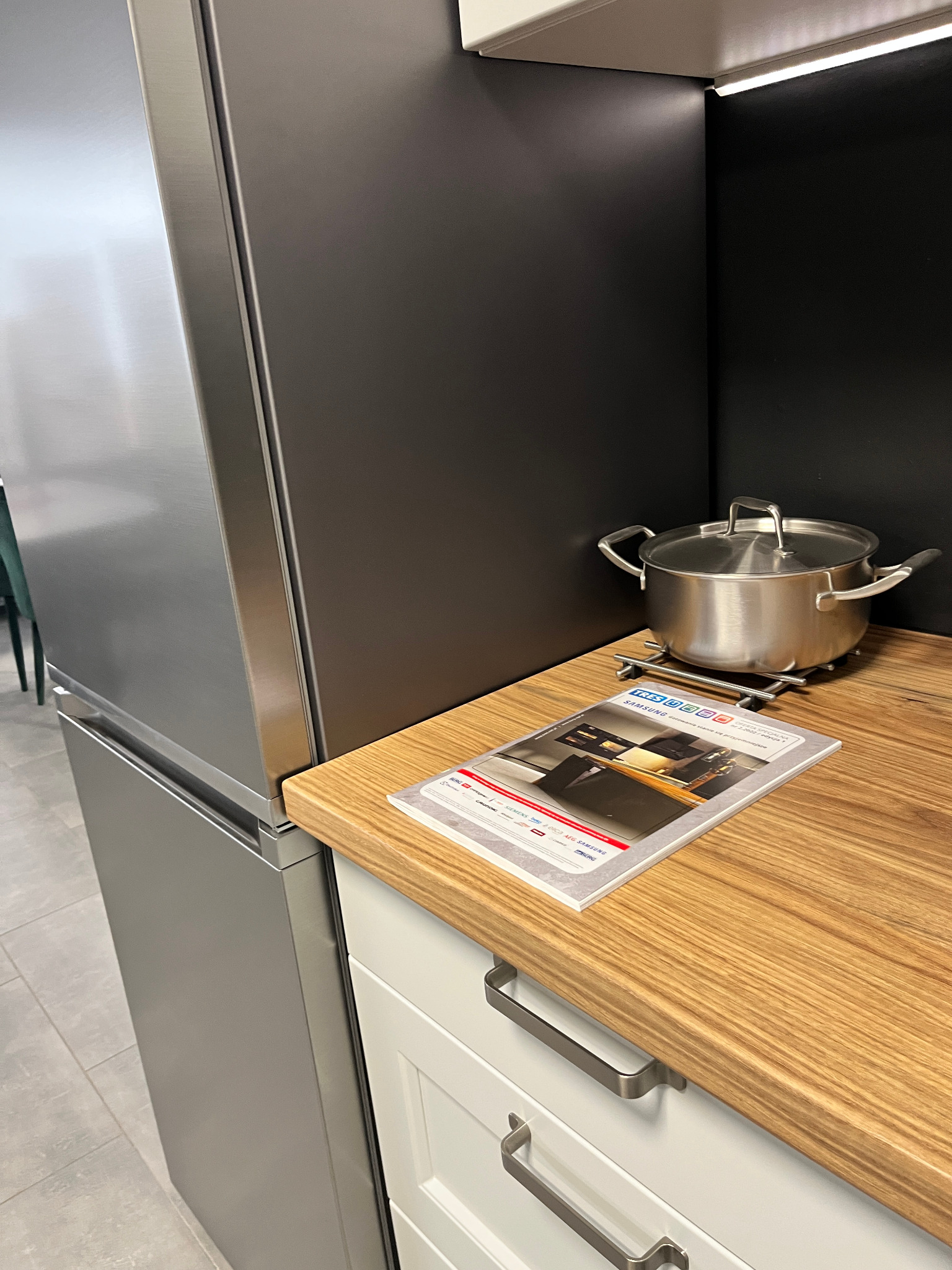 Fragment nowoczesnej kuchni: blat drewniany z garnkiem na podkładce i katalogiem sprzętu AGD, obok lodówka i szafki w zabudowie.