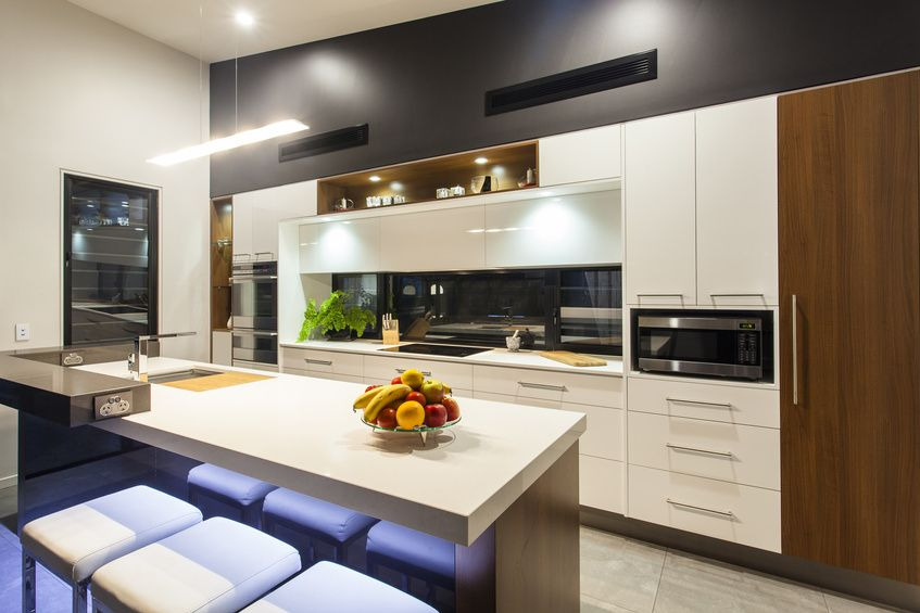 Nowoczesna kuchnia z białymi szafkami, drewnianymi akcentami i wyspą z podświetlanymi stołkami barowymi. Na blacie wyspy misa z owocami. Ciemnoszara ściana z wbudowanymi kratkami wentylacyjnymi.