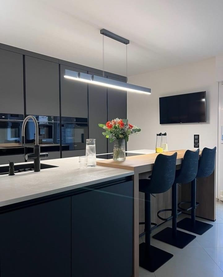 Nowoczesna kuchnia z czarnymi, lakierowanymi frontami szafek, jasnym blatem, drewnianą wyspą z trzema hokerami i wiszącą lampą liniową.