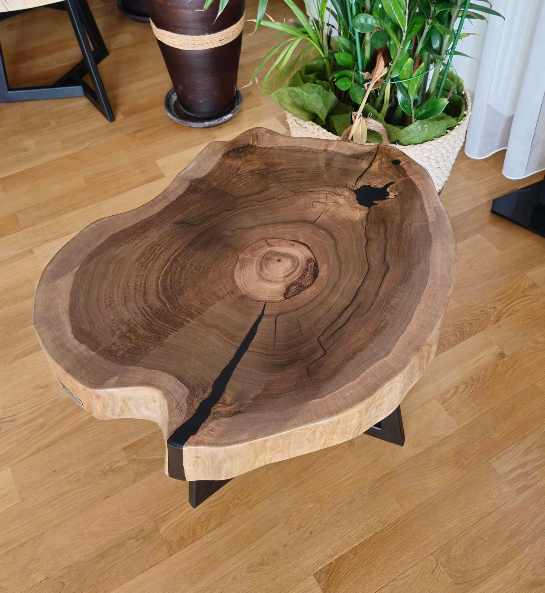 Stolik kawowy z blatem z pojedynczego, nieregularnego plastra drewna z czarną żywicą wypełniającą pęknięcia i ubytki, na metalowych nogach, na podłodze z paneli drewnianych, obok doniczki z kwiatami.