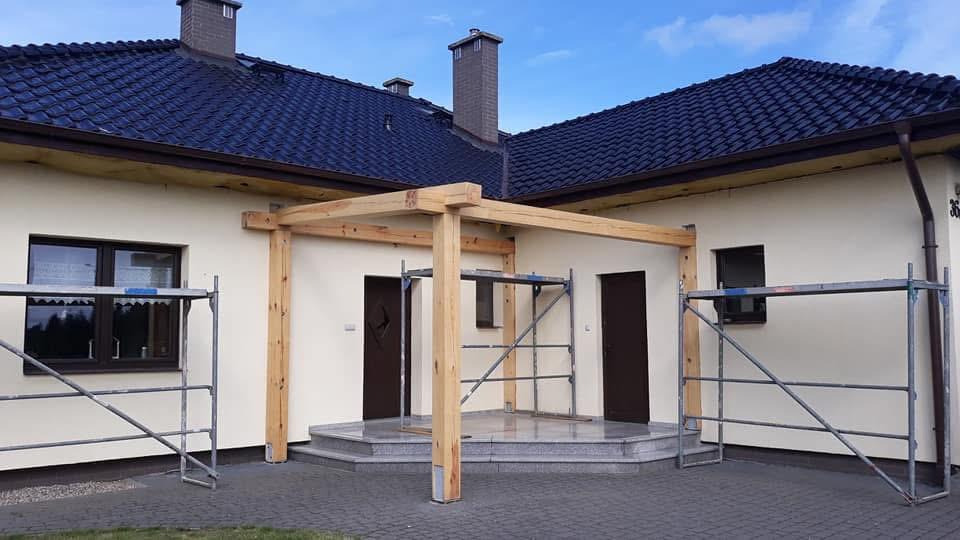 Montaż drewnianej konstrukcji z belek na tarasie domu jednorodzinnego, widoczne rusztowania i wejście na podest.