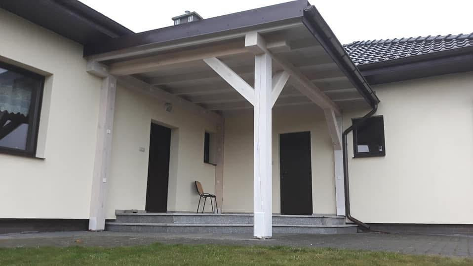 Zadaszone wejście do domu z drewnianą konstrukcją wspartą na białych słupach, schody wyłożone szarymi płytami, widok na dwie pary drzwi wejściowych i krzesło stojące obok.