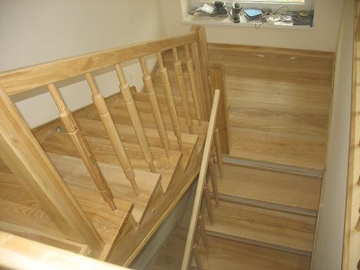 Jasne, drewniane schody z balustradą w naturalnym odcieniu, prowadzące w dół; widok z góry, w tle okno z różnymi przedmiotami.