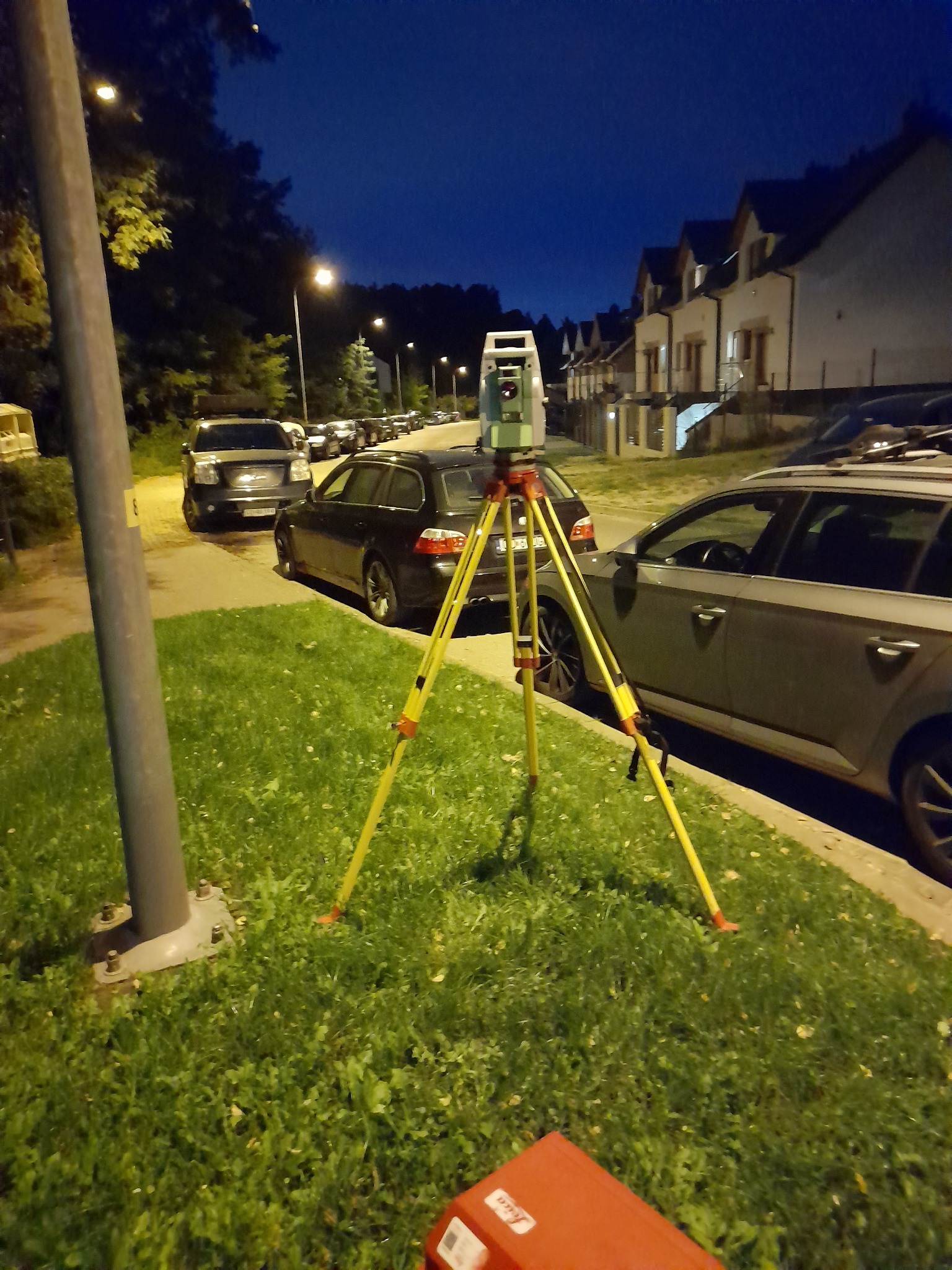Niwelator na trójnogu, ustawiony na trawniku przy ulicy z zaparkowanymi autami i domami w tle, oświetlony światłem latarni ulicznej.