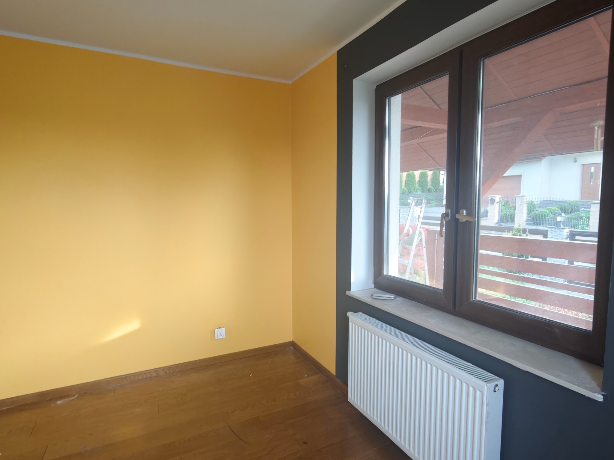 Pokój po malowaniu: żółte ściany, drewniana podłoga, okno z widokiem na dom. Kontrastowe obramowanie okna w kolorze antracytowym. Grzejnik pod oknem.