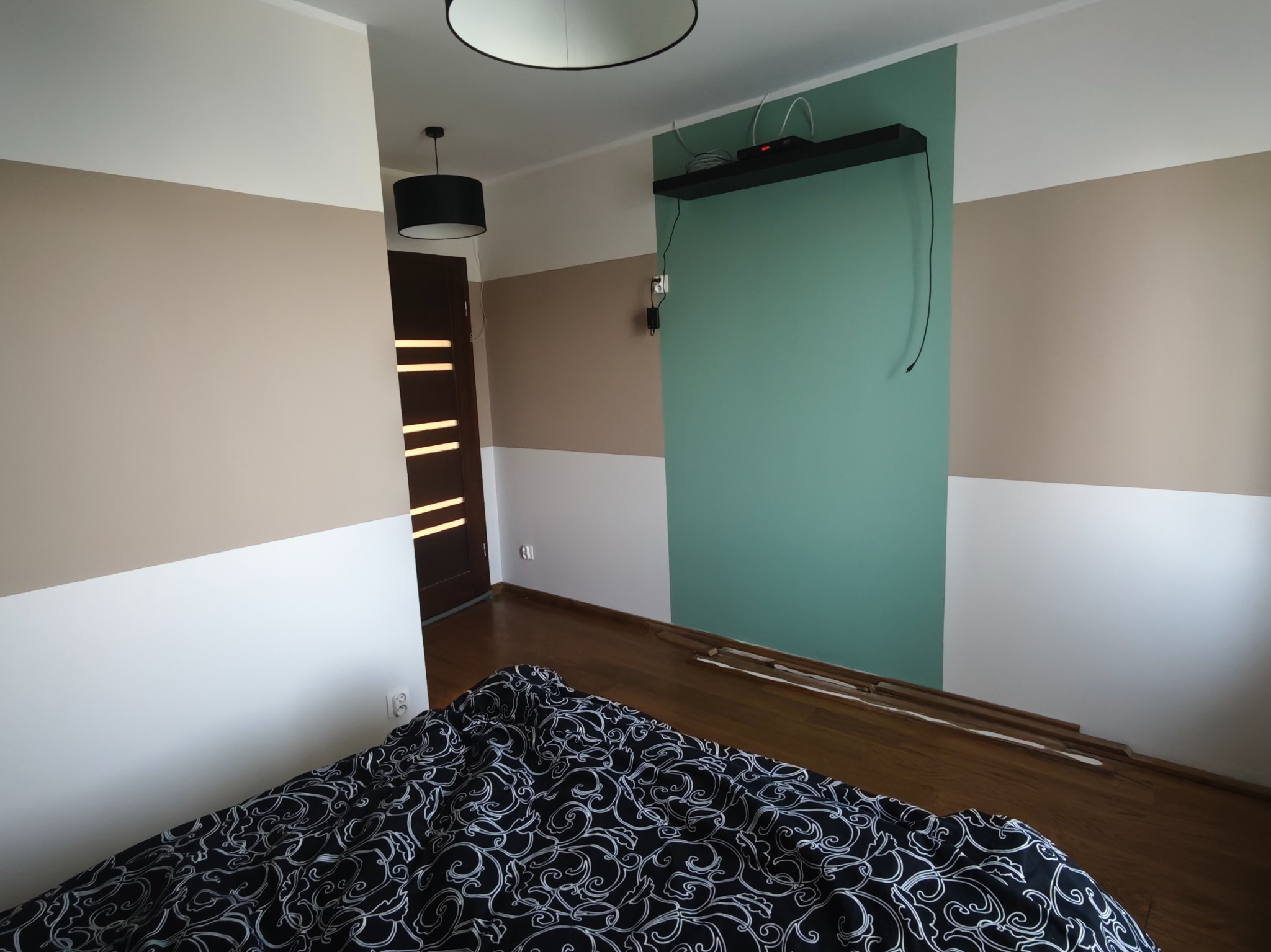 Pokój po malowaniu ścian w pasy: biały, beżowy i miętowy. Widoczne panele podłogowe i fragment łóżka z czarno-białą pościelą. Ciemne drzwi z poziomymi przeszkleniami.