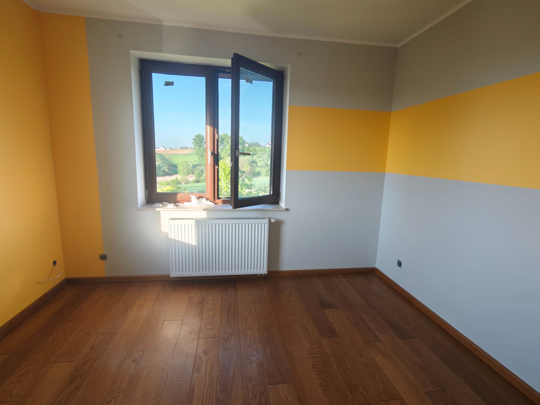 Pokój po malowaniu w Wieliczce: ściany szaro-żółte, drewniana podłoga, otwarte okno z widokiem na zieleń. Nowoczesne wykończenie wnętrz.