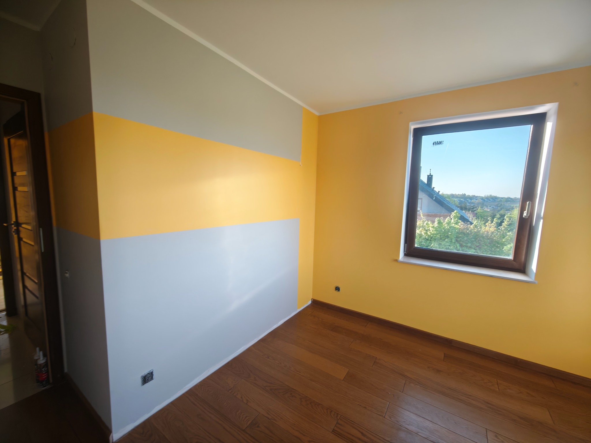 Pokój po malowaniu w pasy: szary, żółty i biały. Parkiet na podłodze, okno z widokiem na zieleń. Widoczne detale wykończenia.